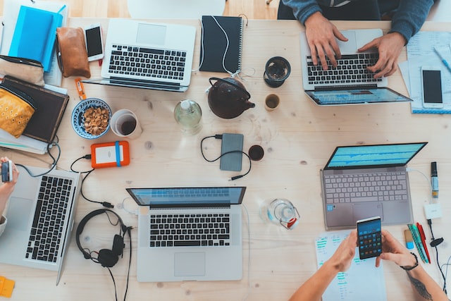 image of a white wooden table with 5 open macs, some phones on it, a couple of power banks and some coffe cups. You can see two hands working on one of the computer and two hands woriking on a phone.