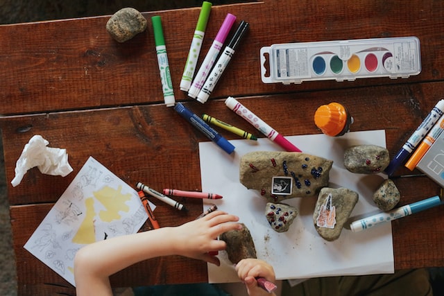 Picture of a brown woodel table with a variety of pencils, markers, papers and some stones on it. You can see two kid hands doodling on a stone