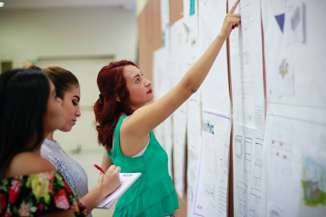 image of 3 women wokring on something. A lot of documents hanging on a wall. One of the women is pointing at something, one is taking notes and the third is looking at the document the first women is pointing at
